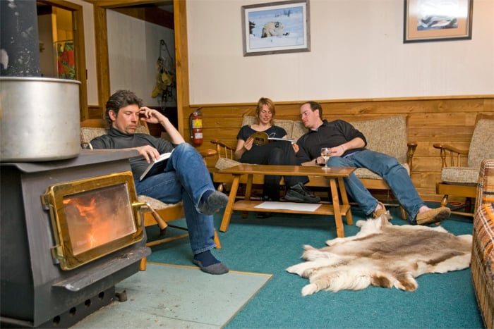 Interior of the lodge showing a common area with couches, chairs, three people reading, and a cozy woodstove